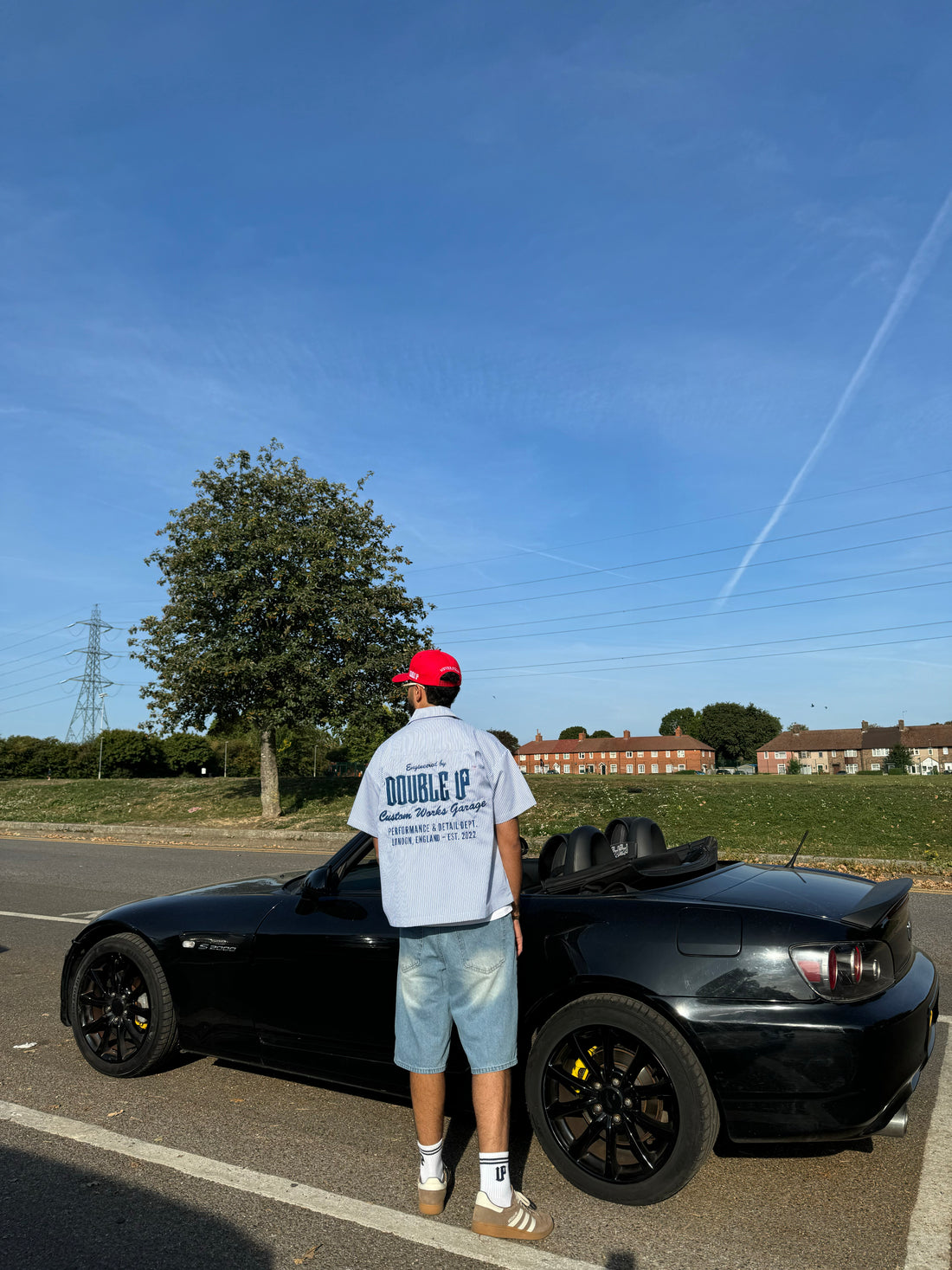 Person standing next to a black convertible car on a road with a clear blue sky.
