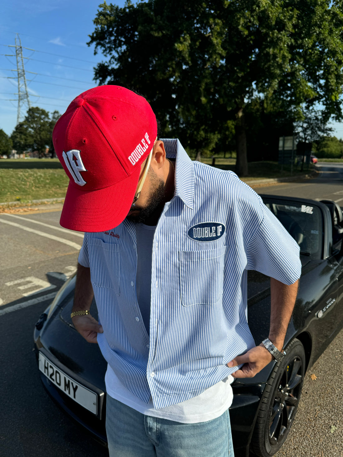 Man wearing a red cap and light blue shirt with a logo, standing next to a black car.